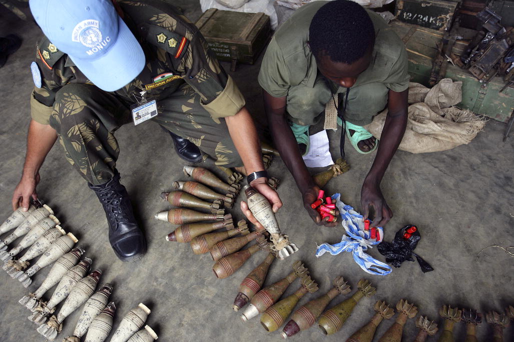 Un homme en treillis et une casquette bleue de l'ONU s'agenouille et fait le point sur les armes tandis qu'un autre homme sans chapeau aide.