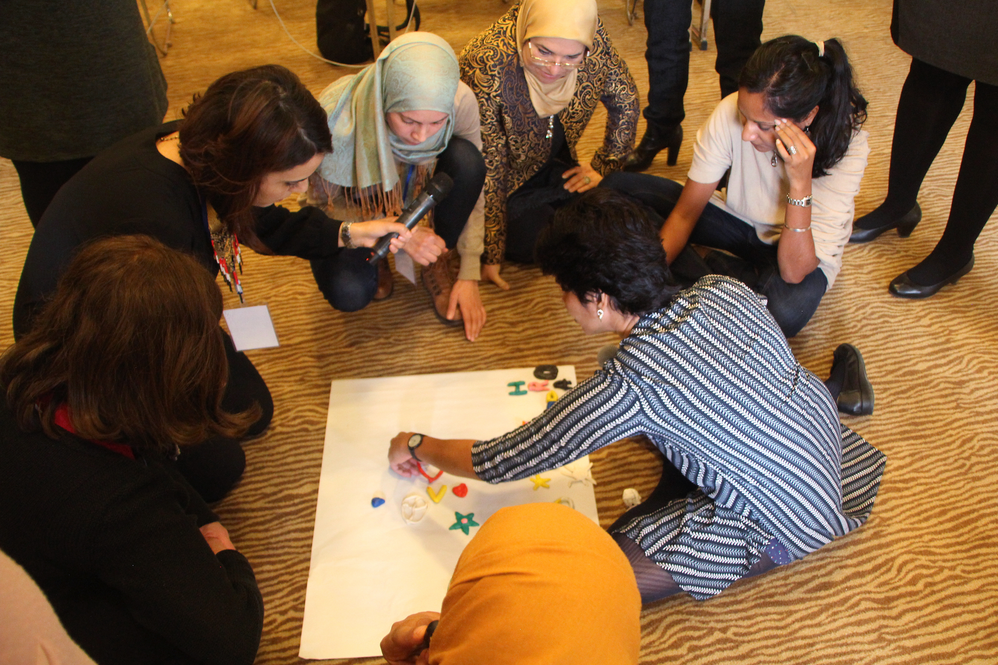 Women kneel on the floor participating in a communications building workshop, one woman holds a microphone out to another woman so she can speak.