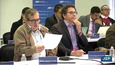 Men sit in a room, two men are in the foreground sitting at a table in what appears to be a hearing, holding up papers.