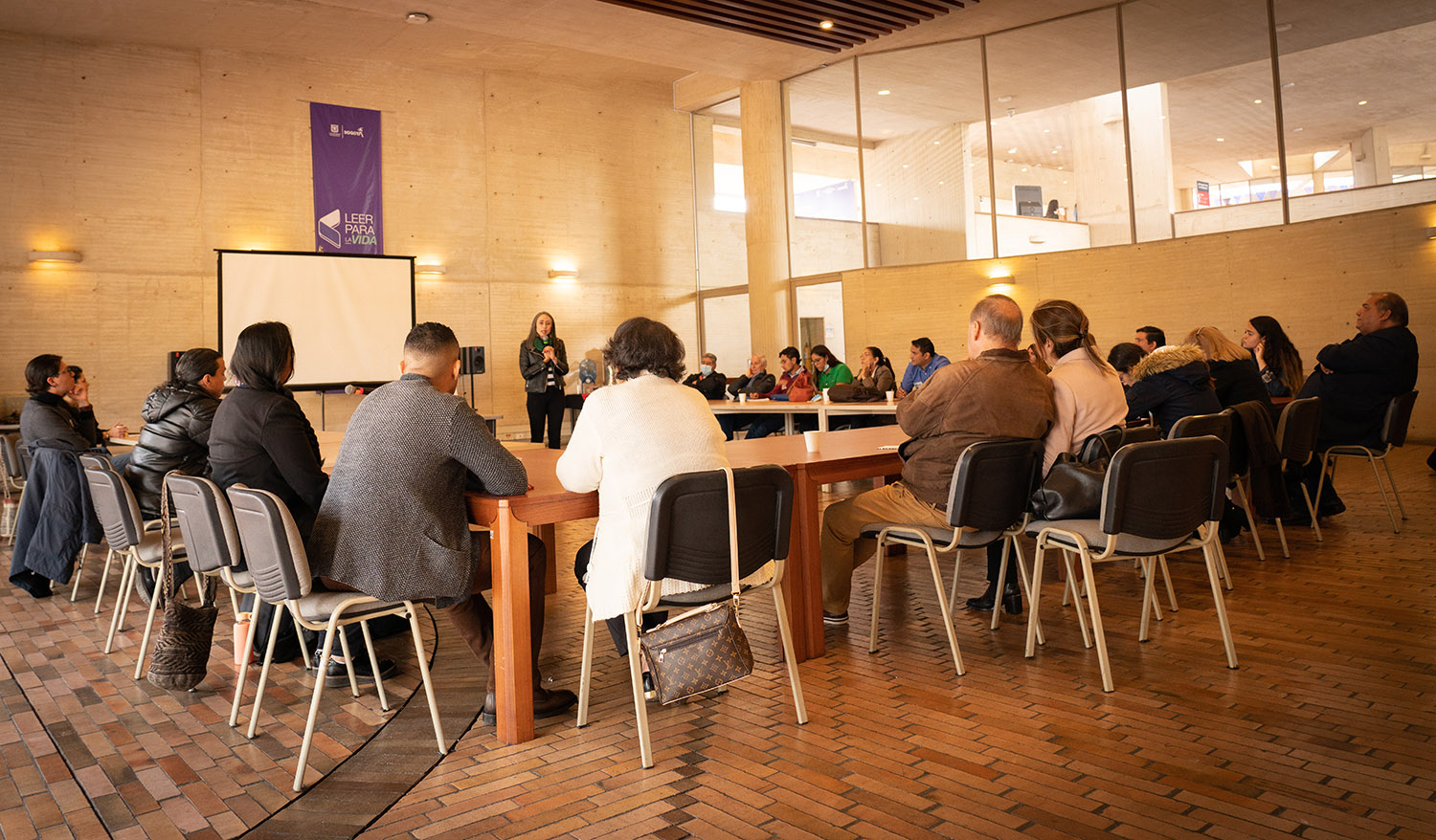 In a large room, a group of people sit around a large table in a semi circle, listening to a woman speaking in the center