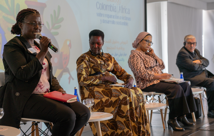 Three women and one man are seated on a stage. One woman is speaking in a microphone.