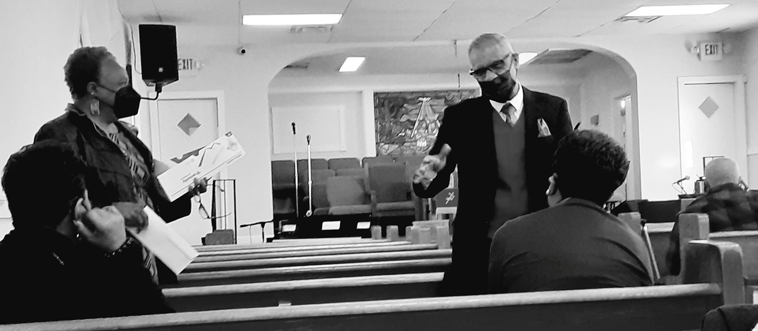 In black and white, a man talks to three people seated in a church pew. 