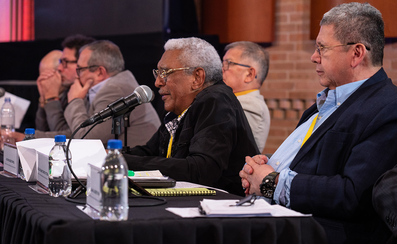 Several men sit at a table, one man leans forward, speaking into a microphone.