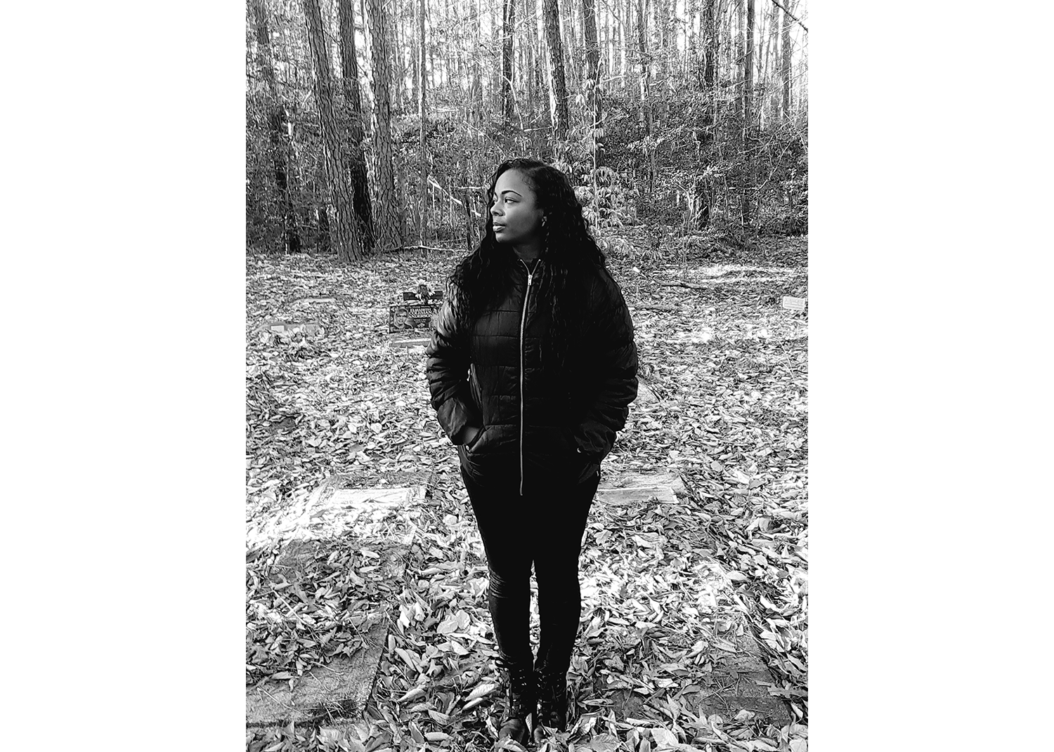 In black and white, a Black woman stands with her hands in her jacket pockets, looking to the left of the frame, in a cemetery with grass and trees around her.