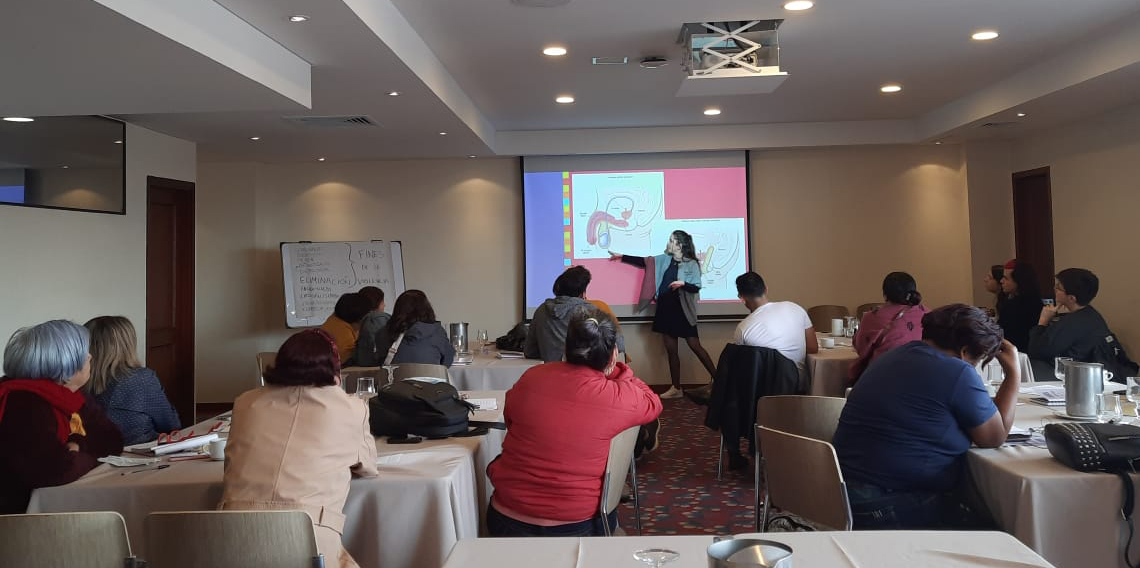 People are seated at tables looking at a person giving a presentation at the front of the room.