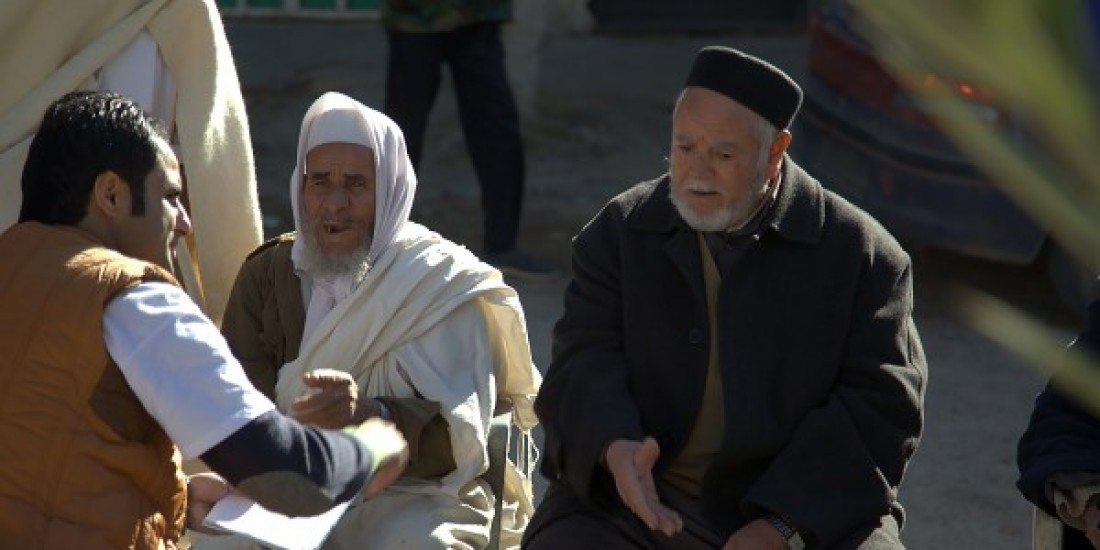 There are three men in discussion with each other, the man on the left in an orange vest is holding a piece of paper and listening to the other two men. 