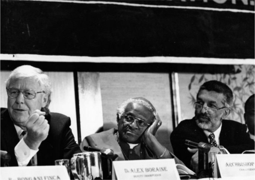 Three men sit behind a desk with name placards in front of them. The name placard that is legible says, "Alex Boraine."