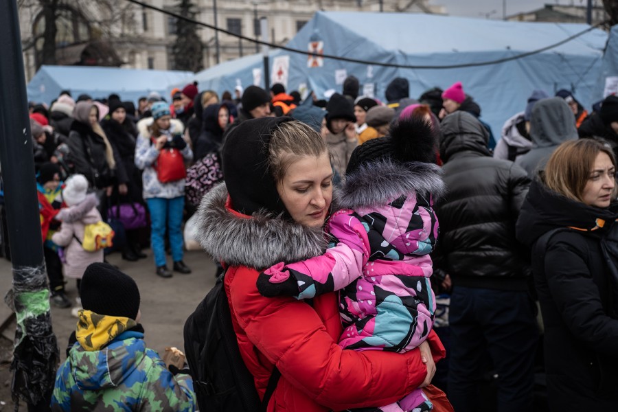 A woman holds a child in a crowd of people around humanitarian assistance tents.