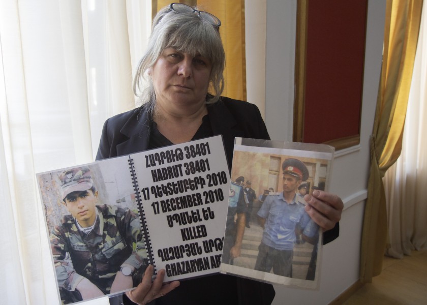 A woman standing indoors holds two large photographs of a young man dressed in military clothing.