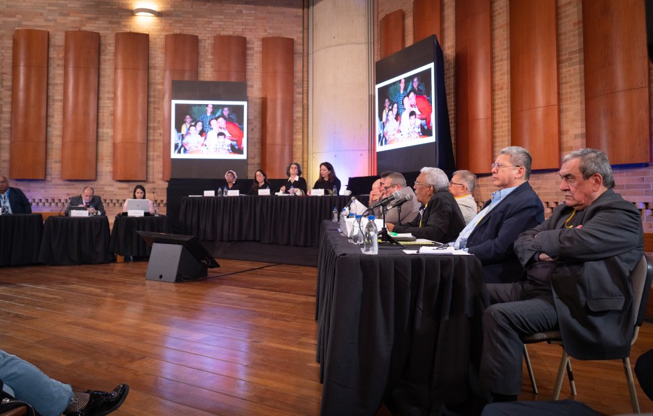 A dozen or so people sit on one side of two long tables all facing the same direction toward others in a room. Above them hang large photographs of a family.