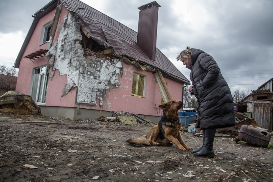 A woman stands near a house whose roof and wall have been severely damaged. 