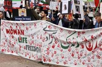 A crowd stands in protest holding a large banner with "Women, Life, Freedom" in front of themselves.