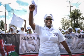 A woman leads a march down a street. Behind her are a banner and other marchers.